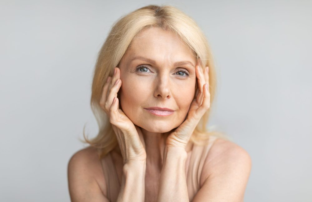 Portrait of beautiful mature blonde woman touching her face, posing over grey studio background. Sensual senior lady with soft skin looking and smiling at camera