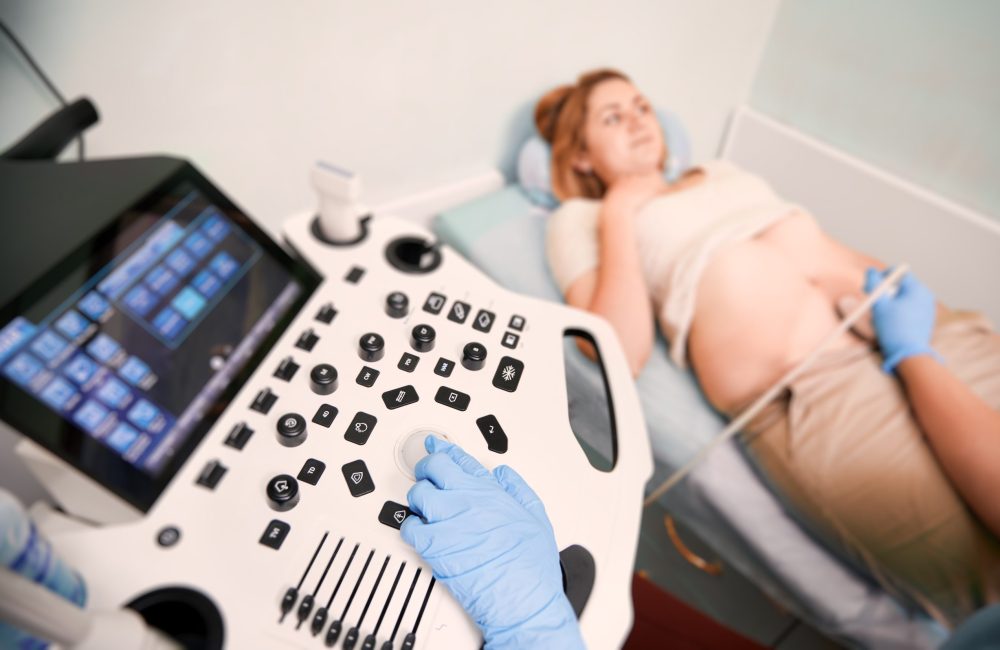 Focus on sonographer's hand in sterile gloves performing ultrasound scanning in clinic. Woman lying on daybed while doctor examining patient abdomen with ultrasound scanner on blurred background.